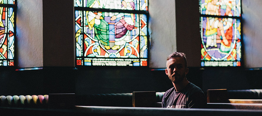 young man sitting in a church pew with stained-glass windows behind